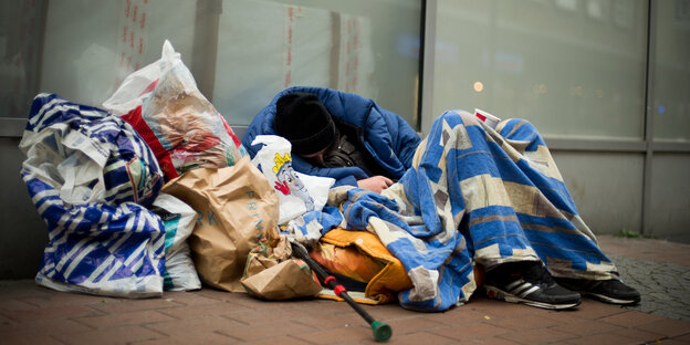 Ein Mann sitzt, eingehüllt in Decken und eine Jacke und umgeben von Tüten auf der Straße
