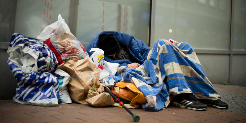 Ein Mann sitzt, eingehüllt in Decken und eine Jacke und umgeben von Tüten auf der Straße