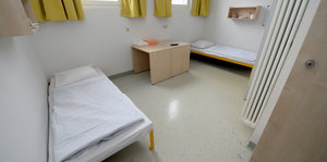 Eine Gefängniszelle. An den Wänden stehen sich gegenüber zwei Betten, in der Mitte ein kleiner Tisch