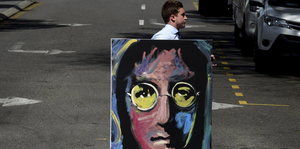 Ein Mann trägt ein psychedelisch buntes Porträt John Lennons über eine Straße