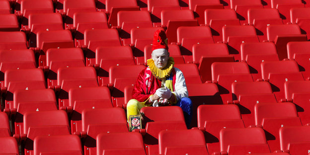 Ein als Clown verkleideter Mann sitzt in einer Reihe aus roten Schalensitzen im Stadion und guckt auf sein Handy