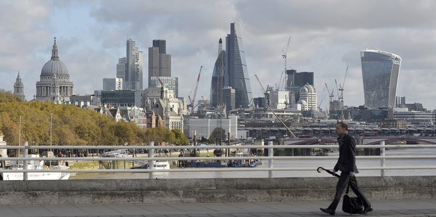 Viele Hochhäuser im Londoner Finanzdistrikt. Davor läuft ein Mann mit Tasche und Regenschirm
