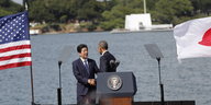 Zwei Männer schütteln sich vor einem Podest die Hände, rechts die japanische, links die US-amerikanische Flagge.