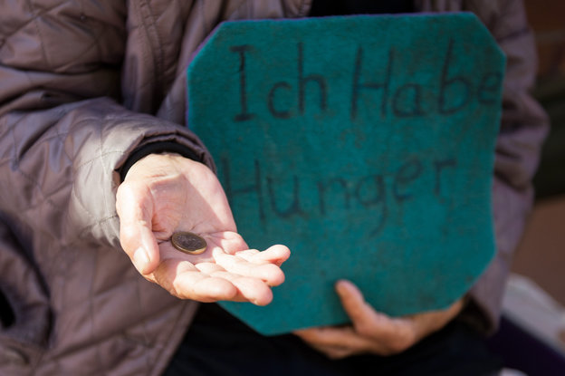 Ausgestreckte Hand mit einem Euro drin im Bild. außerdem trägt die Person ein Schild im Arm, mit der Aufschrift: Ich habe Hunger.