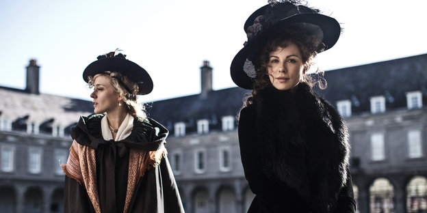 Zwei Frauen in altmodischen Kleidern und mit Hüten vor einem großen englischen Anwesen