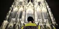Eine Frau mit Polizeiweste ist von hinten zu sehen, wie sie nachts vor dem erleuchteten Kölner Dom steht
