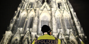 Eine Frau mit Polizeiweste ist von hinten zu sehen, wie sie nachts vor dem erleuchteten Kölner Dom steht