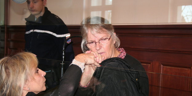 Eine Frau sitzt hinter einer Glasscheibe und blickt zu Boden, eine andere hält ihr tröstend die Hand