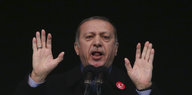 Recep Tayyip Erdogan steht hinter einem Mikrofon, öffnet den Mund und hebt die Hände