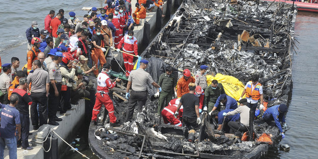 Viele Menschen, teilweise in Rettungsanzügen, steigen von einem abgebrannten Holzboot auf einen Steg