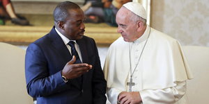 Zwei Männer nebeneinander unterhalten sich, der eine in Papstkleidung, der andere in Anzug und Krawatte