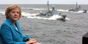 Angela Merkel steht auf einem Schiff mit kleineren Marineschiffen im Hintergrund
