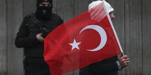 Ein Vermummter mit Maschinengewehr steht an einer Wand. Ein Mann mit türkischer Nationalflagge läuft vor ihm vorbei