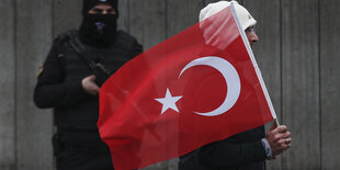 Ein Vermummter mit Maschinengewehr steht an einer Wand. Ein Mann mit türkischer Nationalflagge läuft vor ihm vorbei