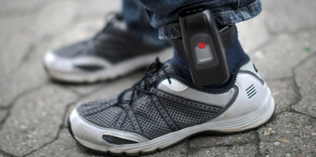 Zwischen Turnschuhen und Jeans eines Mannes ist eine elektronische Fußfessel angebracht