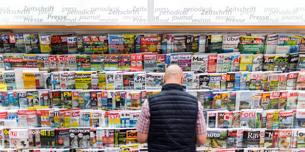 Ein Mann in Weste steht mit dem Rücken zum Bild vor einem Zeitschriftenständer.