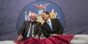 Bernie Sanders und Andrew Cuomo sitzen nebeneinander. Hinter ihnen hängt das Wappen von New York.