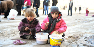 Zwei kleine Mädchen sitzen, in Anoraks gehüllt, vor zwei Eimern mit Essen auf der Straße