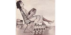 Schwarz-Weiß-Foto: Frau sitzt im Stuhl und hält Magazin in den Händen, schaut dabei in die Kamera. Daneben ein Zeitungsständer
