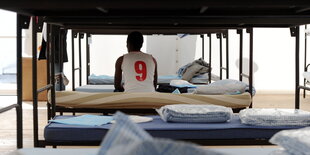 Ein von hinten fotografierter Flüchtling sitzt in einem Unterkunftszelt auf seinem Bett