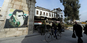 Auf einem Poster an einer Mauer ist Bashar al-Assad abgebildet, auf der Straße davor laufen Menschen