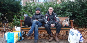 Zwei Obdachlose sitzen, von Tüten begleitet, auf einer Parkbank