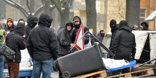 Autonome errichten eine Straßenblockade aus Möbeln und Holz. In der Bildmitte hinter der Blockade stehen zwei vermummte Männer, die sich umamarmen.