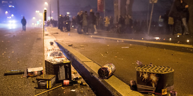 Auf einer Straße liegen Reste von Feuerwerk, dahinter auf dem Gehweg stehen Menschen. Es ist Nacht