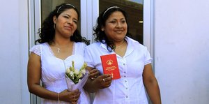Zwei Frauen in weißer Kleidung halten ein rotes Dokument in die Kamera, die eine hält einen Blumenstrauß