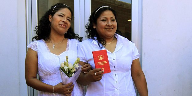 Zwei Frauen in weißer Kleidung halten ein rotes Dokument in die Kamera, die eine hält einen Blumenstrauß