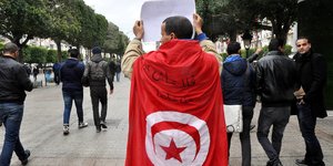 Ein Mann steht allein, umhüllt von einer tunesischen Fahne und hält einen Zettel in die Höhe, vorbeikommende Passanten schauen