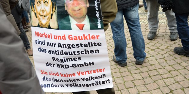 Beinpaare auf einem öffentlichen Platz, dazu ein Plakat, das Merkel und Gauck als Volksverräter beschimpft