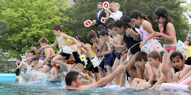 Kinder stehen und sitzen an dem Rand eines Schwimmbeckens