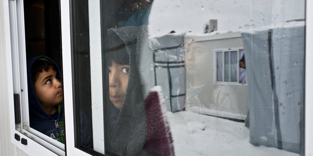 Kinder blicken aus dem Fenster einer Unterkunft, im Fenster spiegeln sich verschneite Wohncontainer