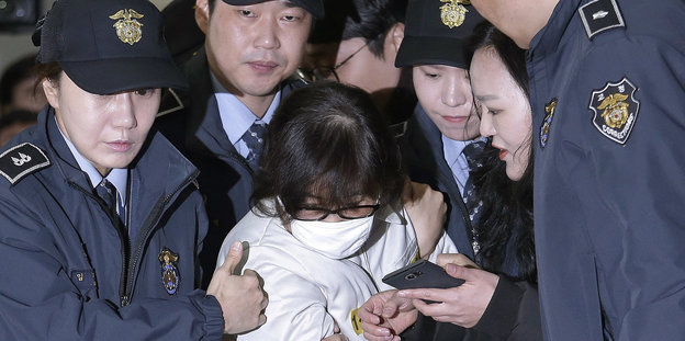 Mehrere Polizist_innen und eine Journalistin umringen eine Frau mit Brille und weißem Mundschutz, die sich duckt