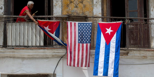 Ein alter Mann beugt sich über die Brüstung eines Balkons. Er hängt drei Flaggen auf. In der Mitte eine us-amerikanische, rechts und links daneben kubanische