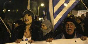 Eine junge und eine ältere Frau sowie ein Mann demonstrieren unter griechischer Flagge