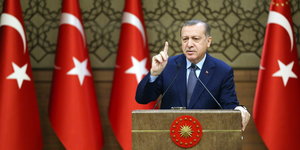 Ein Mann in Anzug steht hinter einem Rednerpult und streckt den Zeigefinger in die Luft. Rechts und links von ihm türkische Flaggen