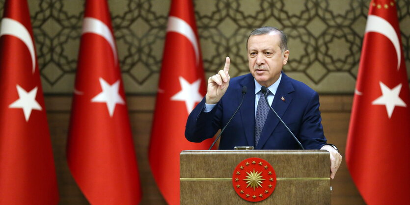 Ein Mann in Anzug steht hinter einem Rednerpult und streckt den Zeigefinger in die Luft. Rechts und links von ihm türkische Flaggen