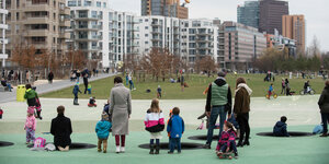 Auf einem grünen Boden stehen Erwachsene und Kinder in bunter Kleidung, dahinter stehen Mehrfamilienhäuser