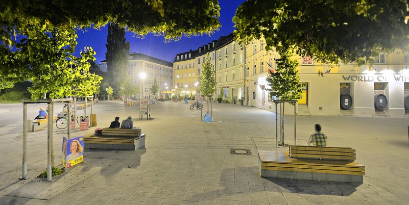 Hellerleuchteter städtischer Platz bei Dunkelheit. Von Bäumen umrahmt, Leute sitzen auf Bänken