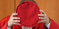 Ein Mann in roter Kutte hält einen roten Hut vors Gesicht