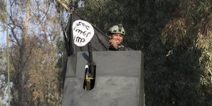 Ein Soldat, der lächelnd aus einem Panzeraufbau hervorschaut, hält eine schwarze Fahne mit einem arabischen Schriftzug in den Händen