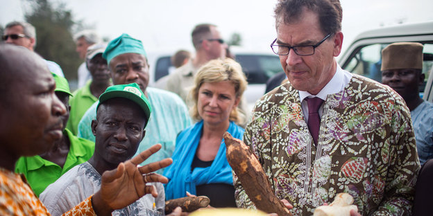 Gerd Müller hält Holzstücke in den Händen und trägt ein buntes Oberteil, neben ihm stehen nigerianische Bauern