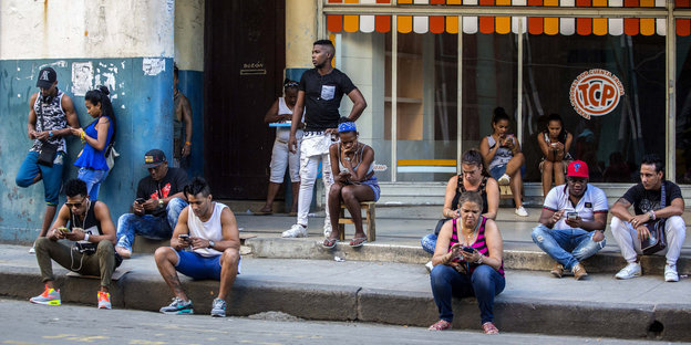 Junge Menschen sitzen vor einem Laden auf dem Gehweg und surfen auf ihren mobilen Engeräten im Internet