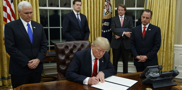 Donald Trump sitzt im Weißen Haus an einem Schreibtisch und unterschreibt etwas, um ihn herum stehen Männer