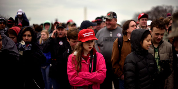 Verschiedene Menschen stehen herum und hören der Amtseinführung von Donald Trump zu, in der Bildmitte: eine junge Frau mit bunter Jacke und rotem Trump-Basecap