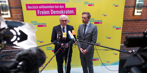 Zwei Männer stehen vor einer gelben Wand. Mikrofone und Kameras sind auf sie gerichtet