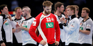 Andreas Wolff steht in einem roten Handballtrikot und mit bedröppelter Miene vor vielen Spielern in weißen Trikots