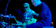 Ein Mann mit Sonnenbrille spielt Schlagzeug, im Vordergrund sitzt ein jüngerer Mann am Laptop, alles ist in blaues Licht getaucht